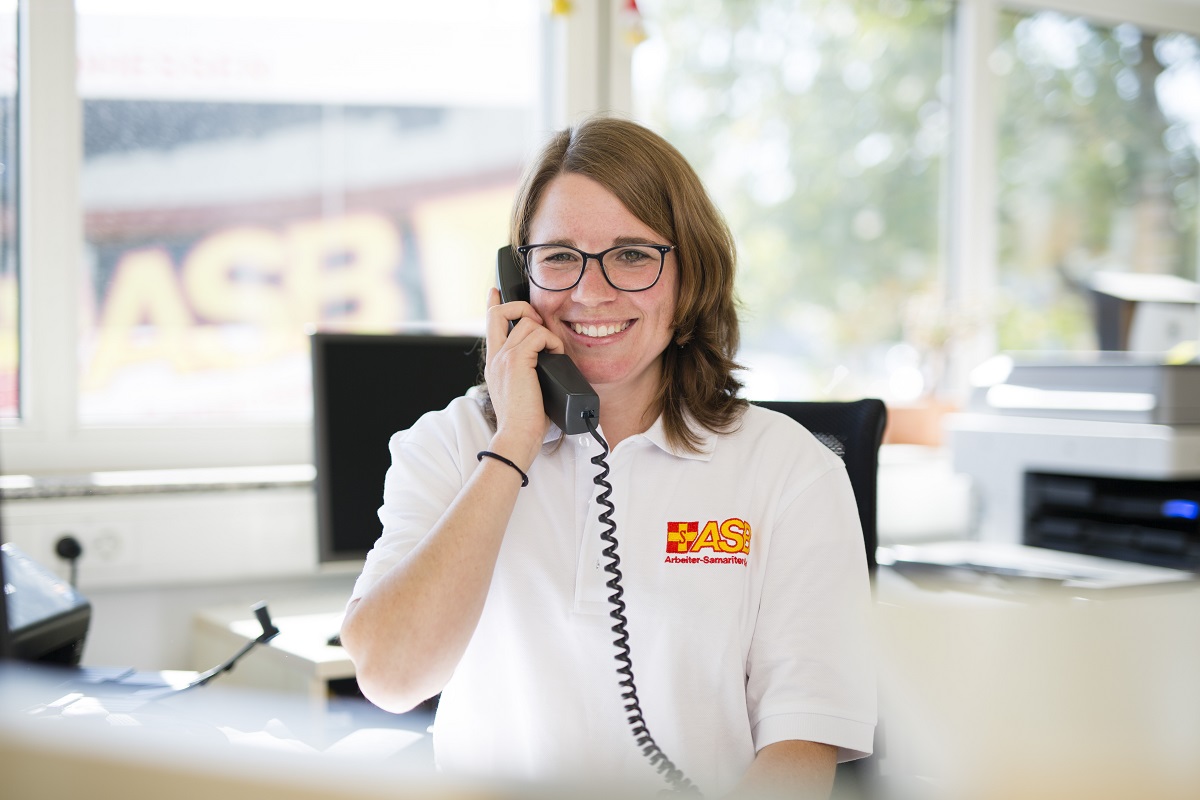 ASB Mitarbeiterin.jpg ASB Mitarbeiterin hält Telefonhörer in der Hand und telefoniert in Büro
