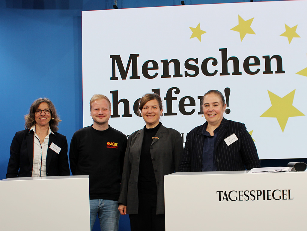 Gruppenfoto vor "Menschen helfen"-Leinwand mit zwei Organisatoriennen und zwei ASB-Vertreter:innen